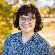 Natasha Riveron Girl with short hair and glasses standing outside smiling