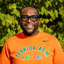 <p>Development Director</p> Man in orange sweatshirt smiling against a green nature background